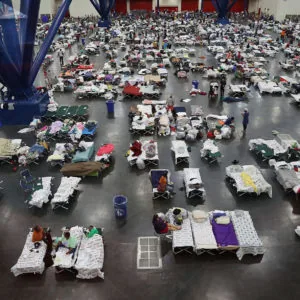 Overhead view of large indoor shelter with hundreds of cots and supplies arranged across a dark floor, with blue structural support beams visible