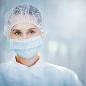 Healthcare worker wearing surgical mask, head covering, and protective gown in medical setting