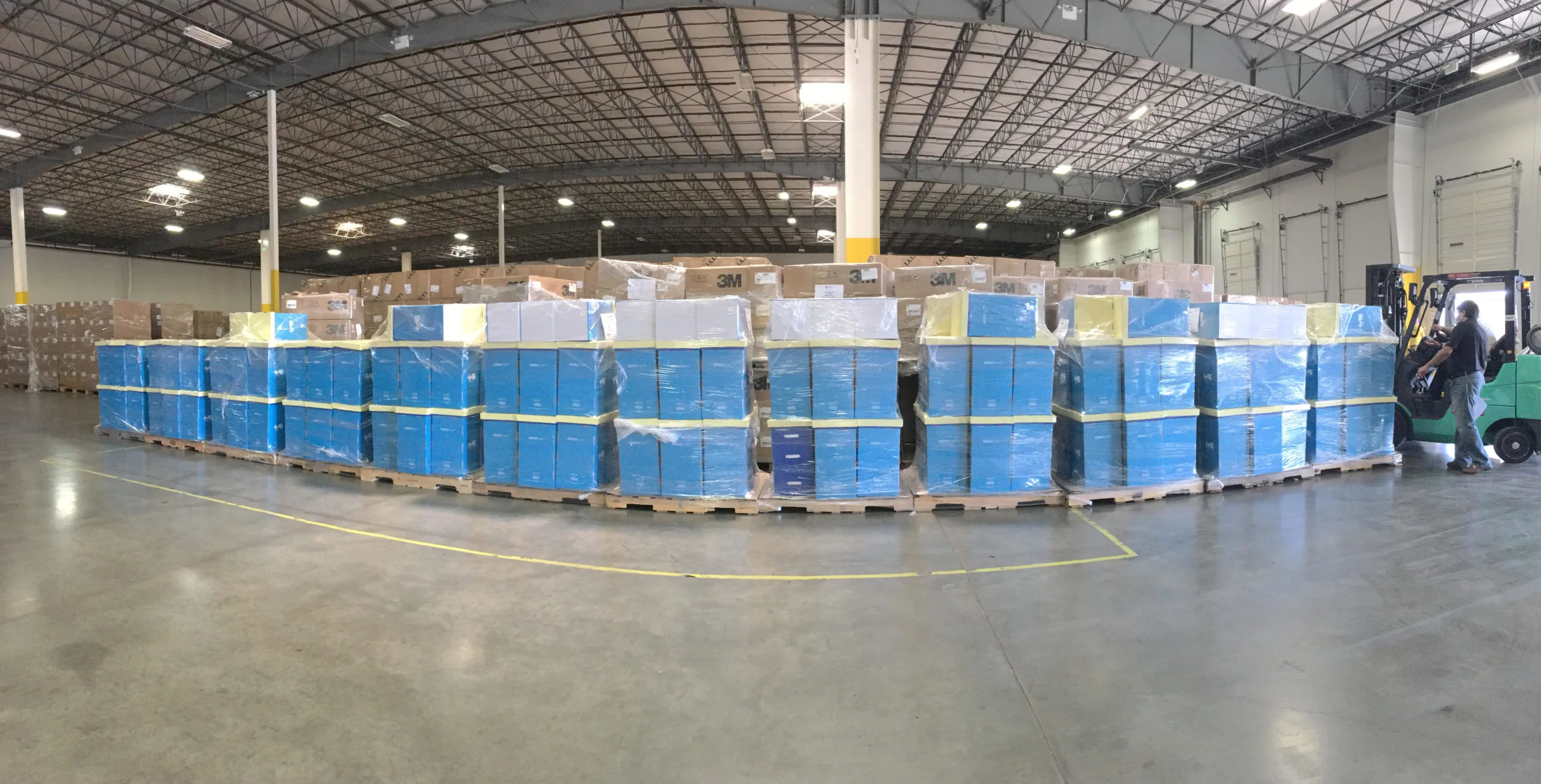 Large warehouse interior with rows of blue plastic totes stacked on wooden pallets, industrial ceiling with lighting, forklift visible on right side