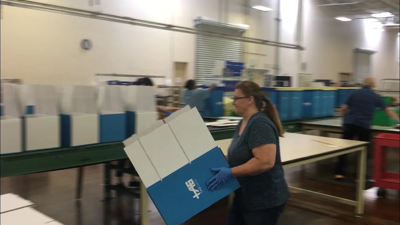 Woman holding white and blue box in industrial warehouse or factory setting with work tables